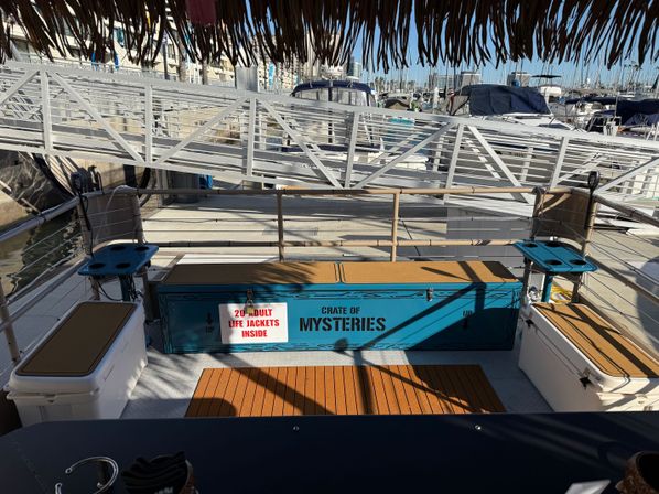 Shaded tiki-roofed boat deck at a marina dock, featuring a bright blue 'Crate of Mysteries' storage chest labeled '20 adult life jackets inside', cushioned benches, cup holders, and docked boats beyond the gangway.