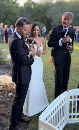 Outdoor wedding on a green lawn: bride in a white gown and groom in a black suit smile while holding adorable puppies, a man in a tux holds another pup beside a white pet playpen with guests blurred in the background.