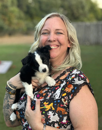 Smiling blonde woman holding a black-and-white puppy in a grassy backyard, wearing a colorful dog-print top and bracelets