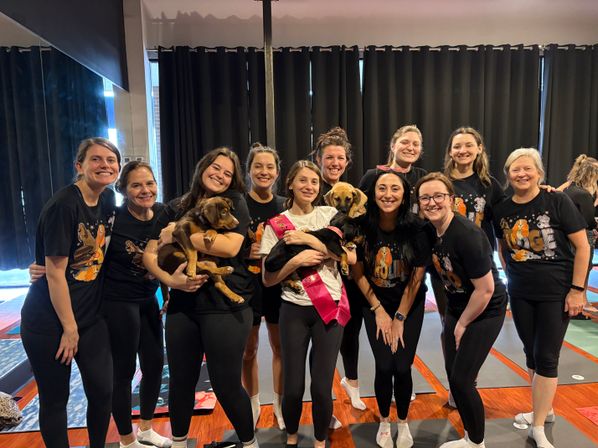 Smiling group of women in a yoga studio wearing matching black tees and leggings, posing on yoga mats while holding playful puppies; one person wears a pink sash for a pup-friendly bachelorette yoga class vibe.