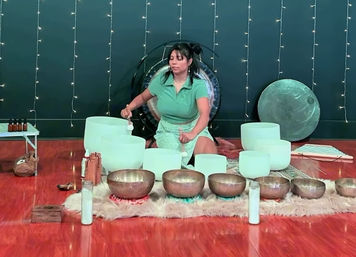 Person leading a calming sound bath in a meditation studio with crystal singing bowls, bronze Tibetan bowls, a gong and twinkling string lights.