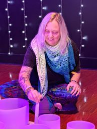 Person seated cross-legged in a cozy wellness studio playing crystal singing bowls with a mallet during a sound bath, purple ambient lighting, string lights, colorful leggings and a blue crocheted shawl.