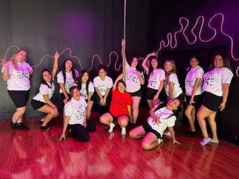 Lively group photo in a neon-lit pole dance studio — women in matching white tops and black shorts with one person in red, posing around a dance pole on a glossy hardwood floor with pink neon wall accents, perfect for a fun dance class or celebration.