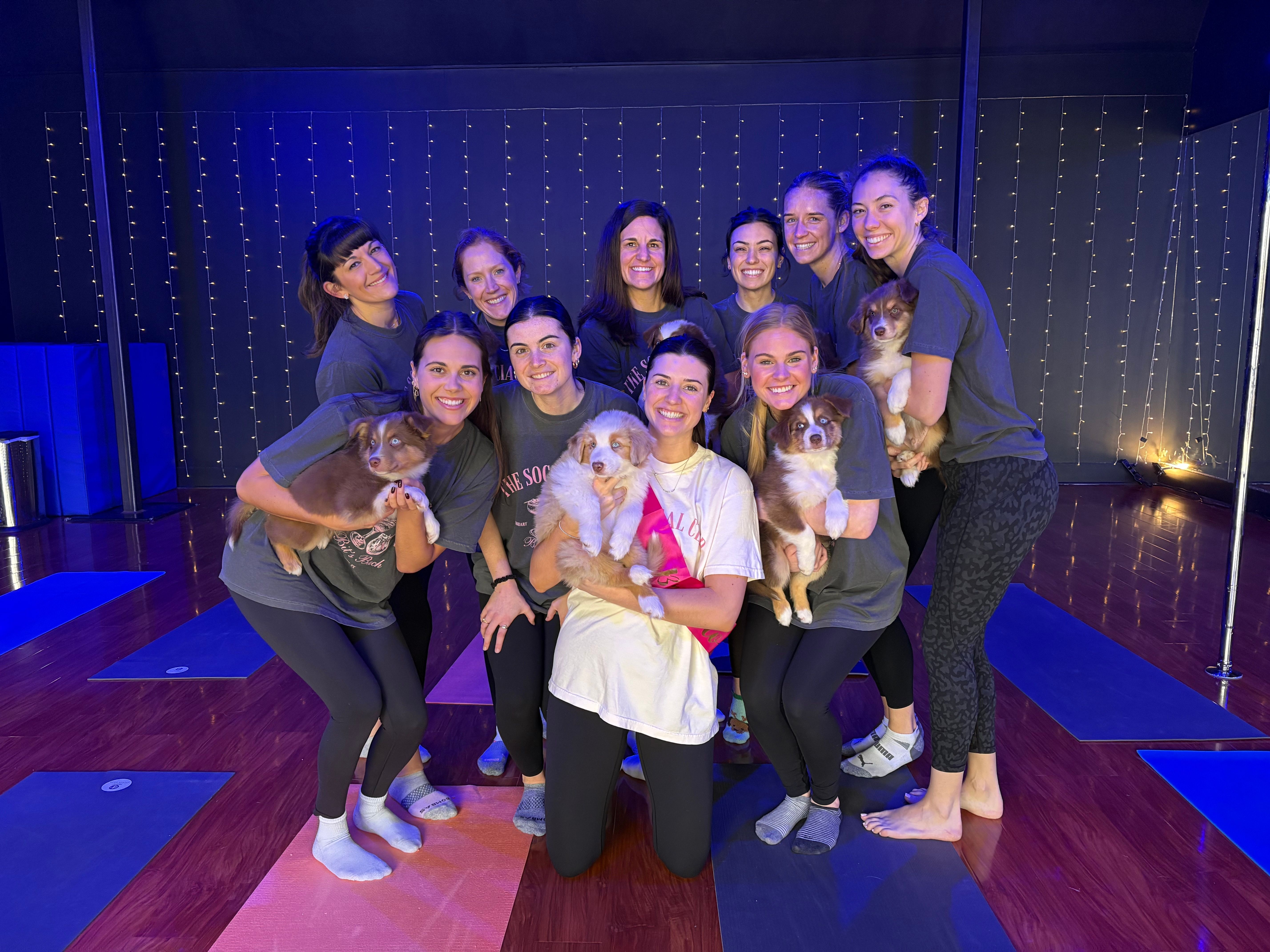 Group of women smiling and holding fluffy brown-and-white puppies during a playful puppy-yoga session on colorful mats in an indoor studio with string lights and a wooden floor.