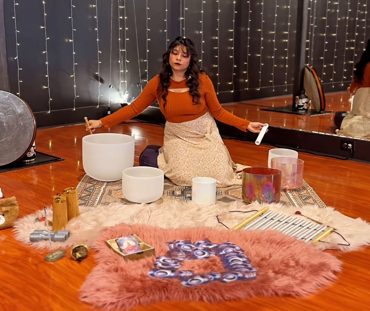 Person seated on rugs in a cozy meditation studio with fairy lights and wooden floor, playing crystal singing bowls and other instruments during a sound bath sound‑healing session.