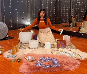 Person seated on rugs in a cozy meditation studio with fairy lights and wooden floor, playing crystal singing bowls and other instruments during a sound bath sound‑healing session.