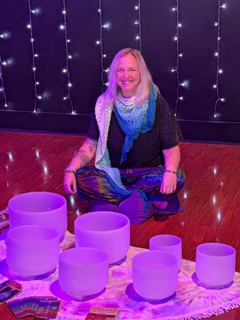 Smiling person seated cross-legged in a cozy purple-lit meditation space with string lights, surrounded by frosted crystal singing bowls on a cloth, wearing a colorful scarf and tie-dye pants with chakra/tarot cards for a sound-healing session.