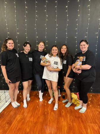Bachelorette party group: six women in coordinating 'bride' and 'babe' shirts smiling and holding two small puppies in an indoor studio with a black wall, vertical fairy lights, and warm hardwood floor.