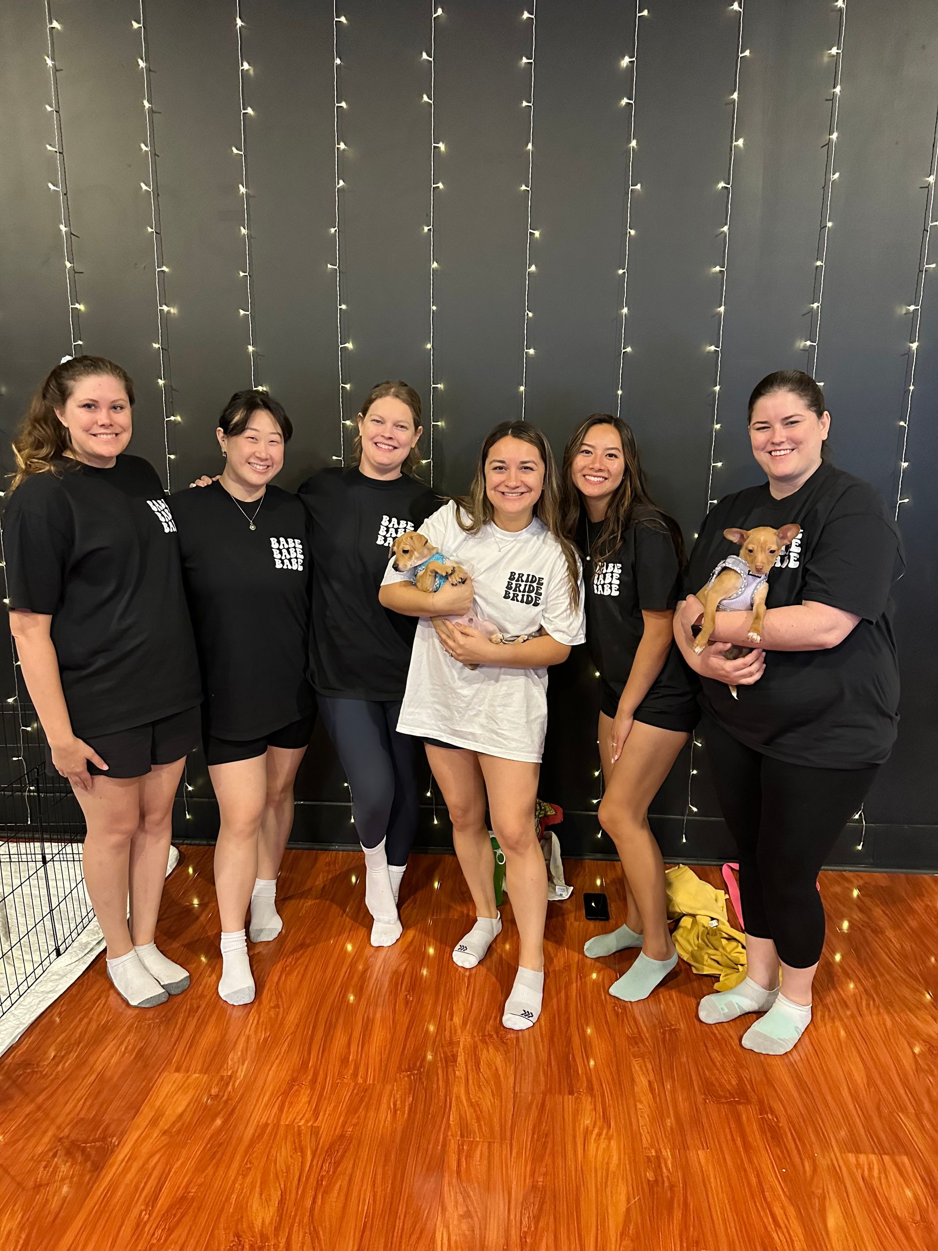 Bachelorette party group: six women in coordinating 'bride' and 'babe' shirts smiling and holding two small puppies in an indoor studio with a black wall, vertical fairy lights, and warm hardwood floor.