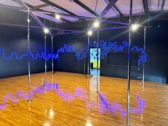 Empty pole fitness studio interior with shiny chrome poles on a glossy hardwood floor, black walls and exposed ceiling beams lit by electric blue neon squiggle lights reflecting across the floor, open doorway to a bright lobby.