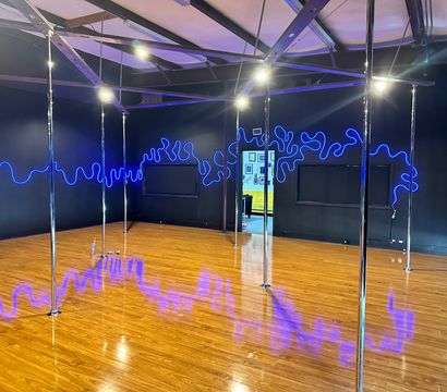 Empty pole fitness studio interior with shiny chrome poles on a glossy hardwood floor, black walls and exposed ceiling beams lit by electric blue neon squiggle lights reflecting across the floor, open doorway to a bright lobby.