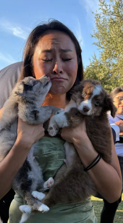 Woman in a green dress holding two fluffy puppies—one nuzzling her cheek, the other staring at the camera—at an outdoor garden party with guests and trees in the background.