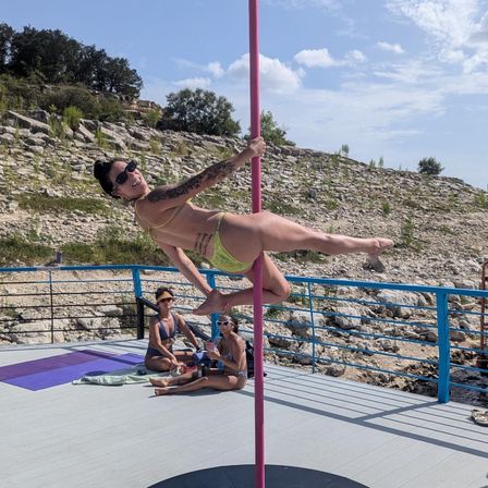 Woman in green bikini holding a horizontal pole-dance pose on a pink pole on a sunny rocky seaside deck, friends lounging nearby