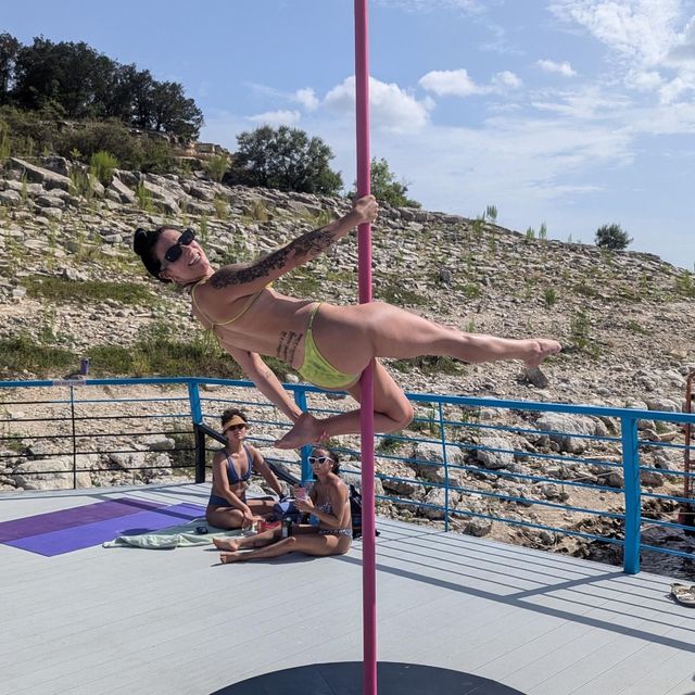Woman in green bikini holding a horizontal pole-dance pose on a pink pole on a sunny rocky seaside deck, friends lounging nearby