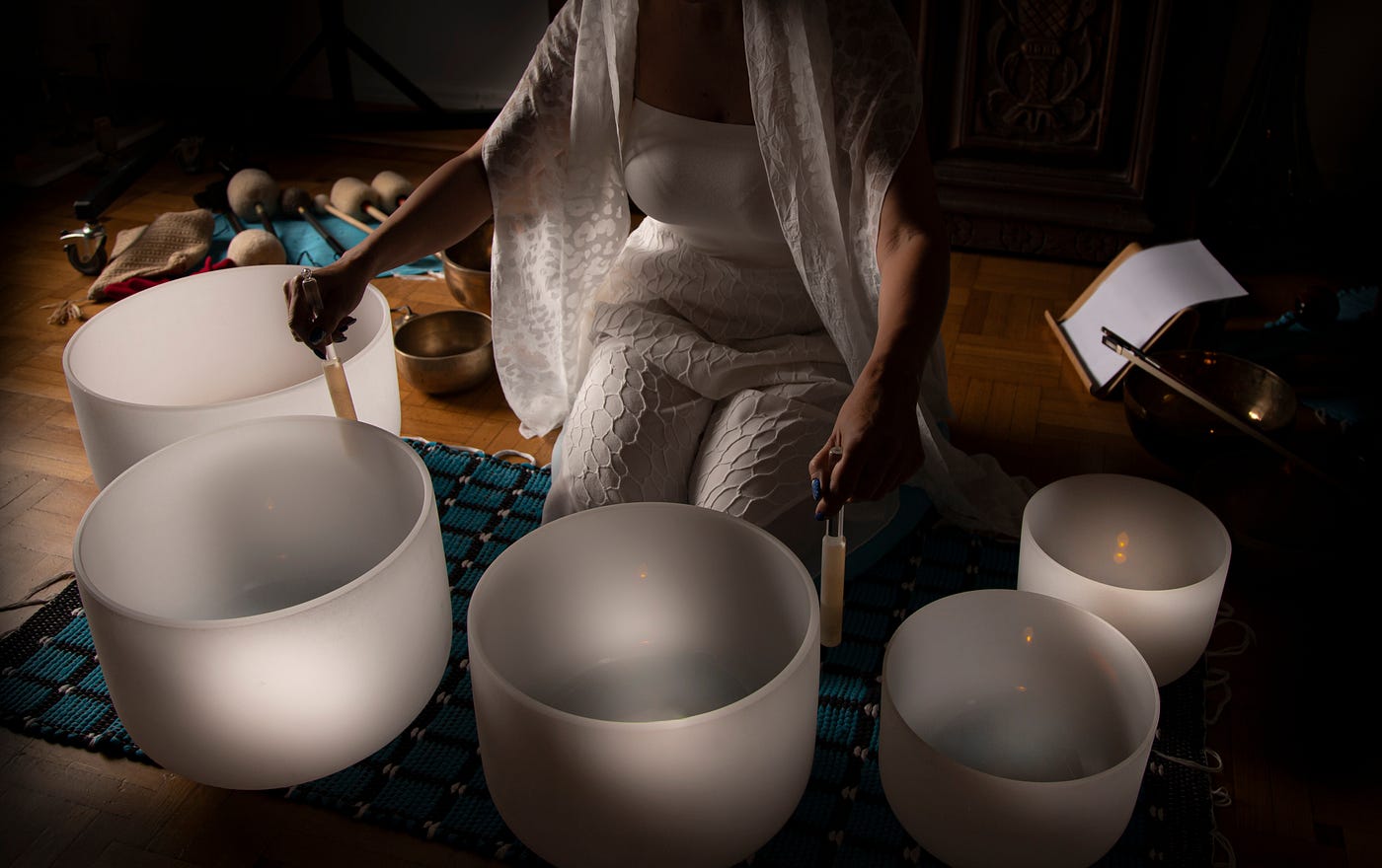 Person in white clothing seated on a woven mat indoors playing frosted crystal singing bowls with mallets for a calming sound-healing meditation session, multiple bowl sizes on a hardwood floor under soft, warm light.