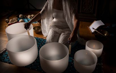 Person in white clothing seated on a woven mat indoors playing frosted crystal singing bowls with mallets for a calming sound-healing meditation session, multiple bowl sizes on a hardwood floor under soft, warm light.
