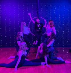 Seven women in an aerial hoop class at a fitness studio, one seated in a suspended hoop while others kneel or do splits on a wood floor under purple-blue stage lighting and string fairy lights.