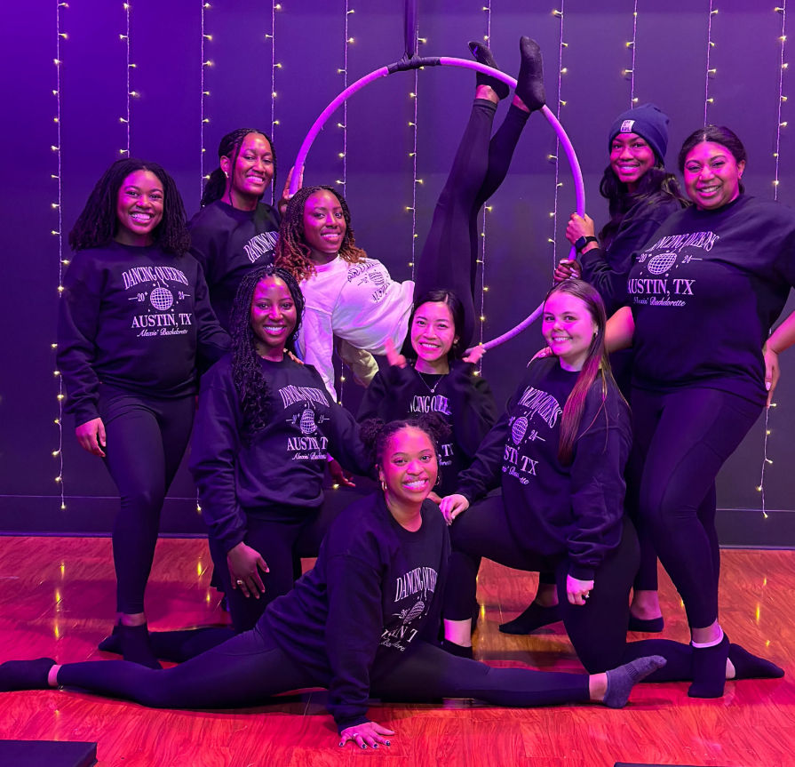 Group of women in matching Austin, TX sweatshirts posing with an aerial hoop (lyra) in a purple-lit fitness studio decorated with string lights