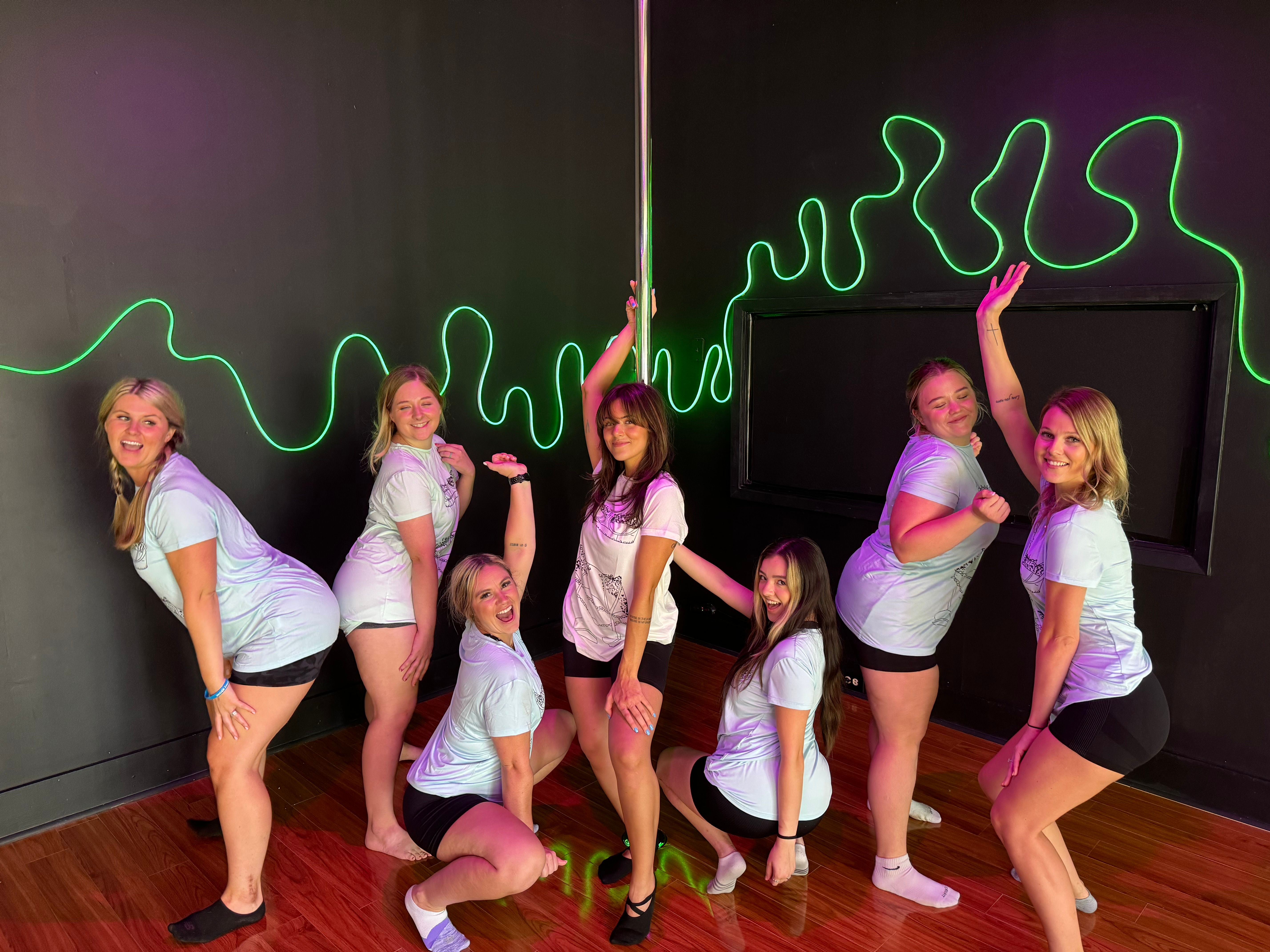 Seven women in matching shirts and shorts posing and smiling around a pole inside a neon-lit dance studio with green squiggle wall art and hardwood floor.