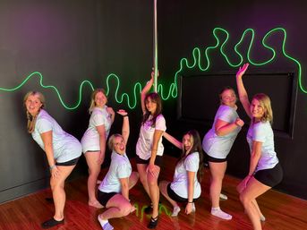 Seven women in matching shirts and shorts posing and smiling around a pole inside a neon-lit dance studio with green squiggle wall art and hardwood floor.