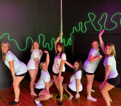 Seven women in matching shirts and shorts posing and smiling around a pole inside a neon-lit dance studio with green squiggle wall art and hardwood floor.
