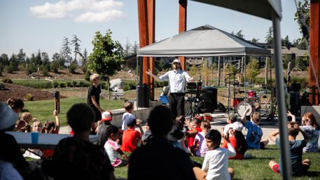 Performer under a canopy on a small outdoor stage engaging a seated group of children on the grass at a sunny community park event, with drums, instruments and trees in the background.