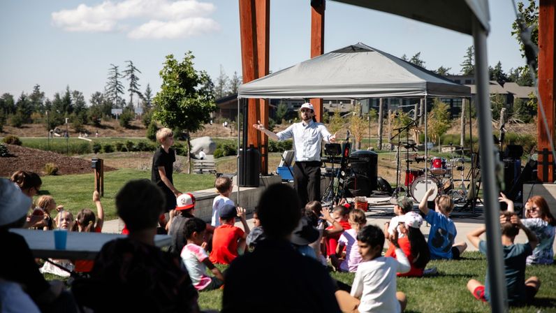 Performer under a canopy on a small outdoor stage engaging a seated group of children on the grass at a sunny community park event, with drums, instruments and trees in the background.