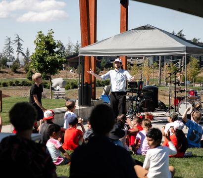 Performer under a canopy on a small outdoor stage engaging a seated group of children on the grass at a sunny community park event, with drums, instruments and trees in the background.