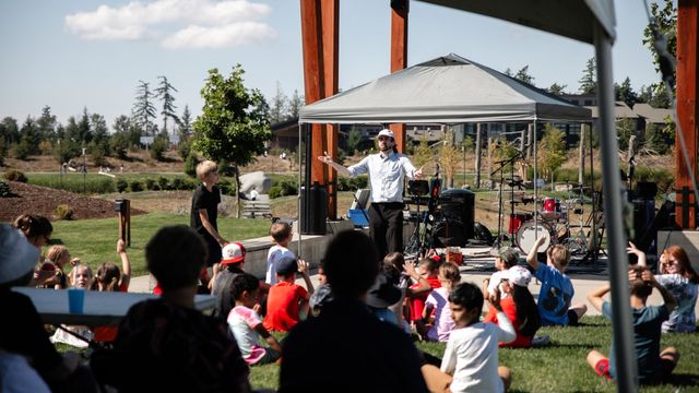 Performer under a canopy on a small outdoor stage engaging a seated group of children on the grass at a sunny community park event, with drums, instruments and trees in the background.