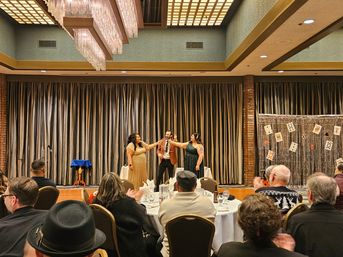 Magician and two women in formal dresses perform a ring trick on a hotel ballroom stage with a playing-card backdrop while guests at round banquet tables applaud.
