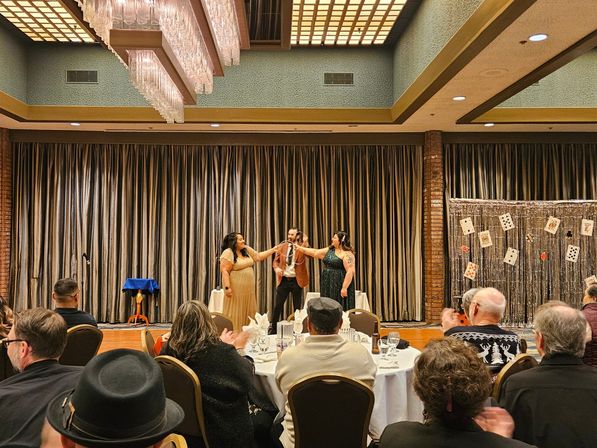 Magician and two women in formal dresses perform a ring trick on a hotel ballroom stage with a playing-card backdrop while guests at round banquet tables applaud.