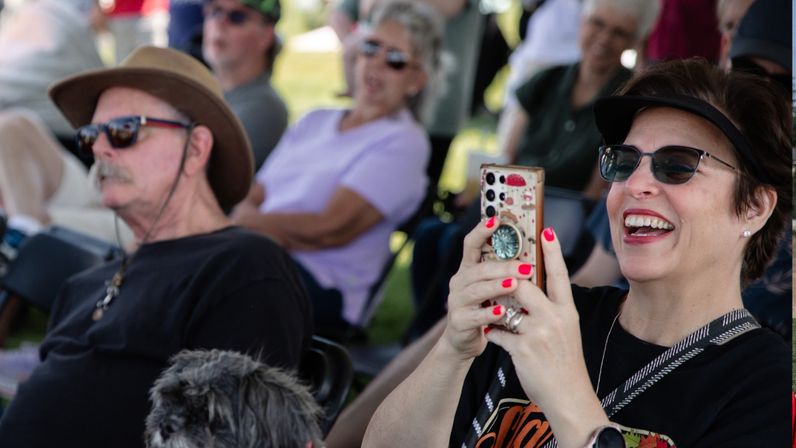 Smiling woman in sunglasses and visor taking a photo with her smartphone at an outdoor park concert, seated beside a man in a hat and a small dog