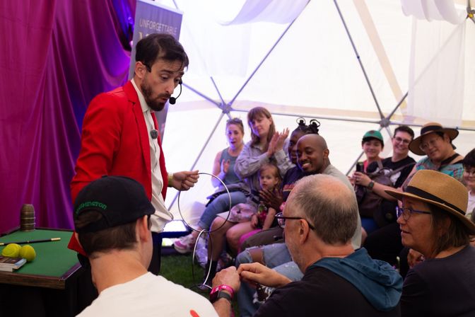 Magician in a red jacket performing a ring trick for a smiling, engaged family audience inside a white festival dome tent with purple curtains