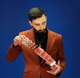 Magician in a rust blazer and black turtleneck performing a cascading spread of red-backed playing cards against a vibrant blue background.