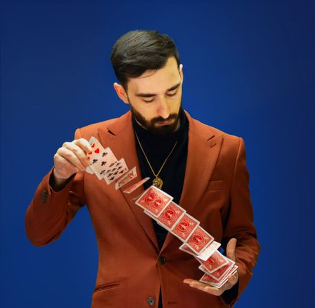 Magician in a rust blazer and black turtleneck performing a cascading spread of red-backed playing cards against a vibrant blue background.