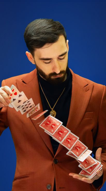 Magician in a rust blazer and black turtleneck performing a cascading spread of red-backed playing cards against a vibrant blue background.