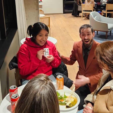 Smiling woman in a red hoodie holding a revealed playing card while a surprised man in a brown suit reacts at a cozy indoor cafe table with drinks and a bowl of food.