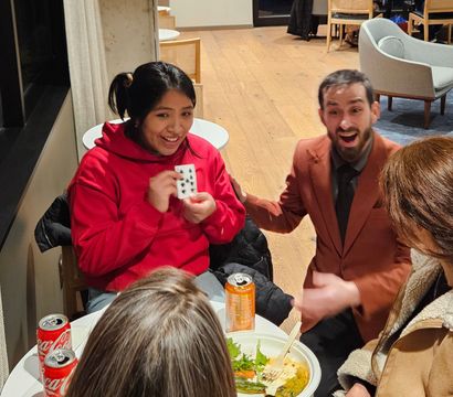 Smiling woman in a red hoodie holding a revealed playing card while a surprised man in a brown suit reacts at a cozy indoor cafe table with drinks and a bowl of food.