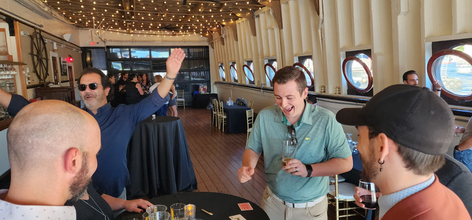 Cheerful group at a waterfront boat party, guests laugh and sip drinks around cocktail tables under string lights aboard a wooden ship with round porthole windows