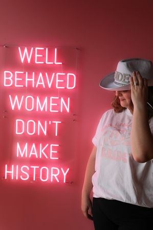 Woman in an embellished white cowboy hat and casual tee poses against a pink backdrop with a bright neon sign reading "WELL BEHAVED WOMEN DON'T MAKE HISTORY"