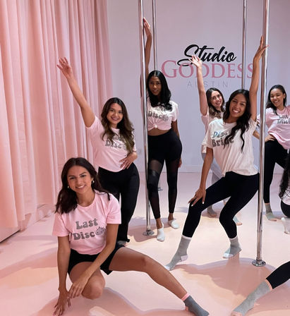 Smiling women striking poses around chrome poles in a pink dance studio, wearing matching 'Last Disco' tees and leggings for a playful bachelorette-style pole fitness class.