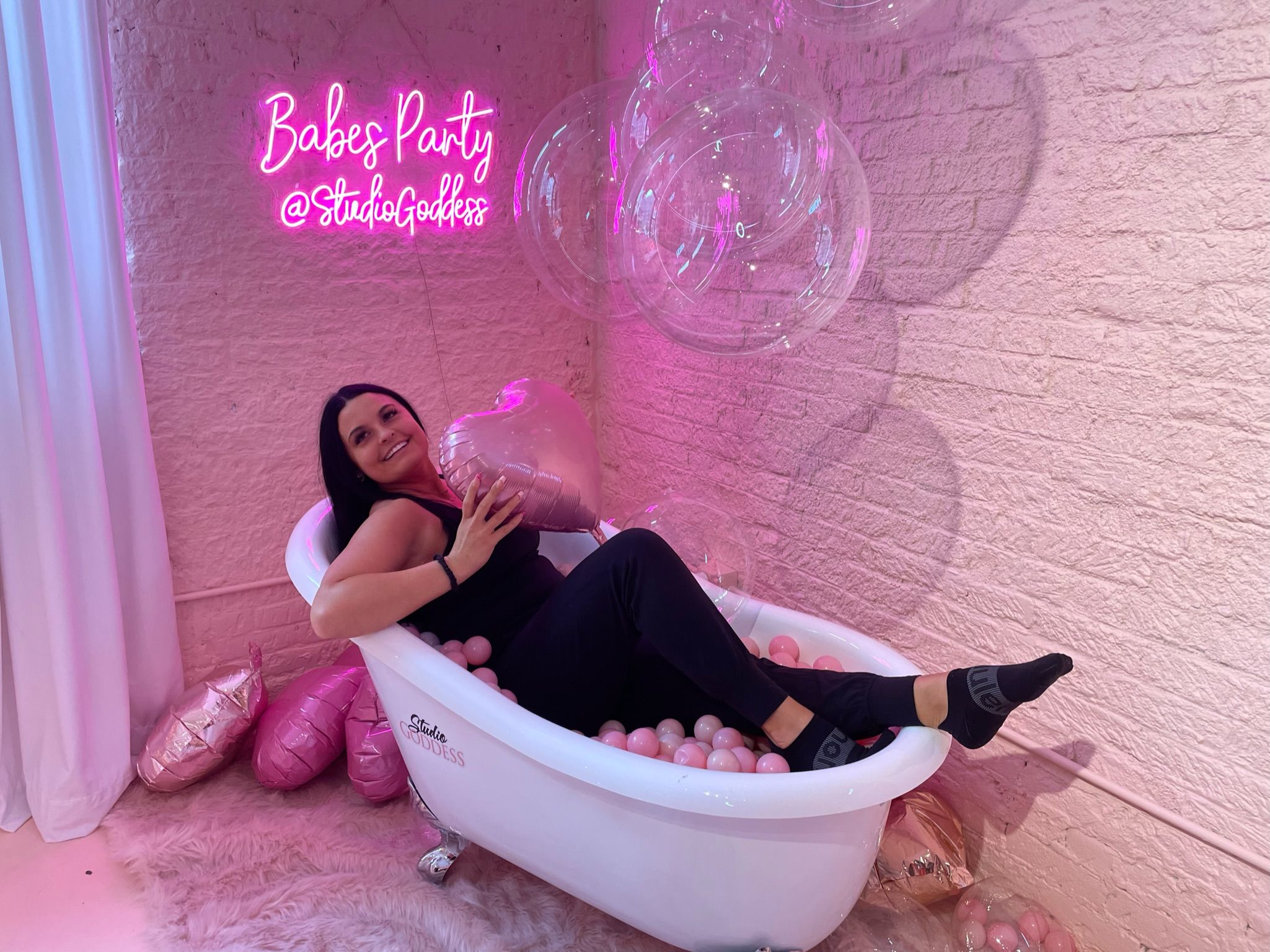 Woman lounging in a white clawfoot bathtub filled with pink ball-pit balls, holding a heart balloon against a pink brick wall with a neon "Babes Party" sign and clear floating balloons — playful indoor party photo setup.
