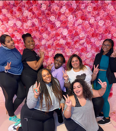 Seven friends smiling and flashing peace signs in casual activewear in front of a vibrant indoor pink rose wall backdrop