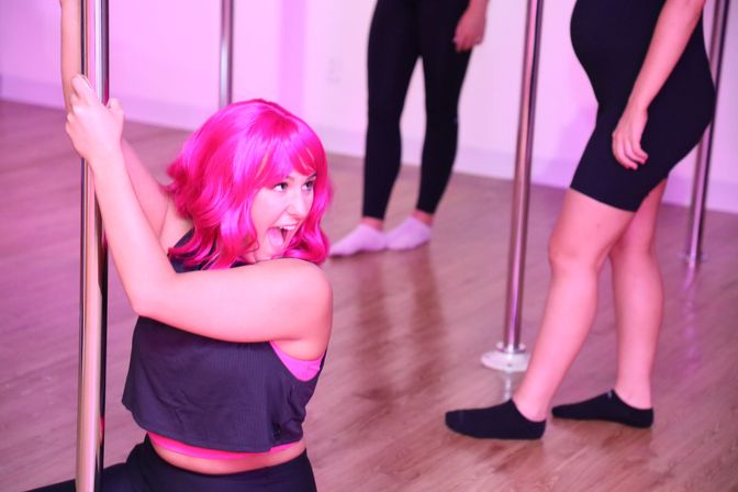 Person with vibrant pink hair laughing while holding a pole in a pink-lit pole dance fitness class, hardwood studio floor and other participants visible