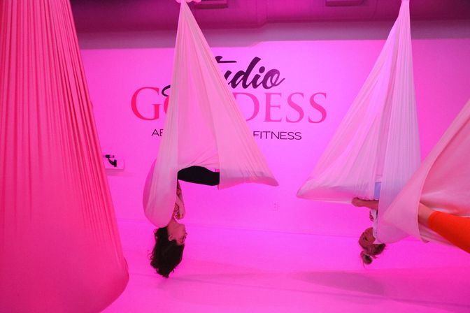Three participants doing inverted poses in white aerial hammocks inside a pink-lit aerial yoga fitness studio.
