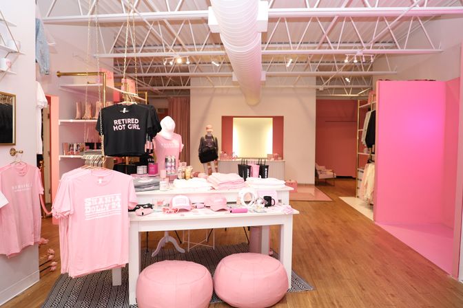 Bright pink women's boutique interior with trendy clothing and accessories on tables and racks, mannequins, round pouf stools, wood floors and exposed white ceiling ductwork