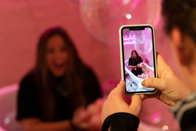 Hands holding a smartphone capturing a laughing woman sitting in a bathtub filled with pink balls against a pink party backdrop with clear bubble decorations.