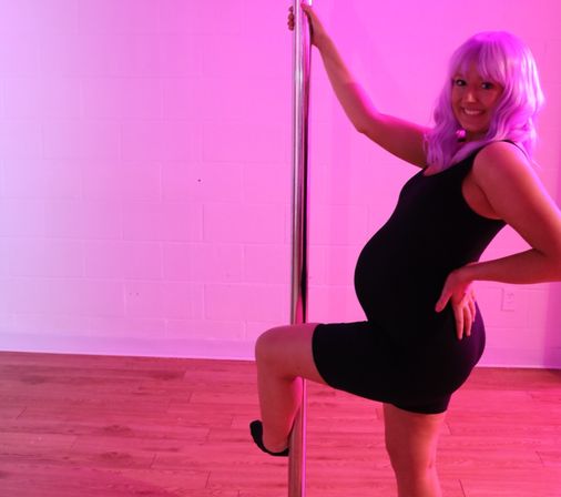 Smiling pregnant pole dancer in a purple wig and black outfit posing in a pink-lit dance studio with a wooden floor