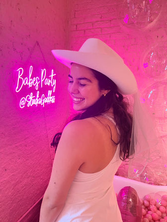 Smiling woman in a white cowgirl hat and veil posing in a pink-lit bachelorette party photo booth with a neon sign, clear balloons and a pink ball pit