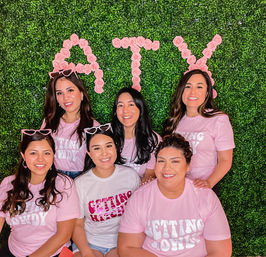 Group photo of six smiling women in matching pink 'Getting' T‑shirts and playful sunglasses, posing in front of a green boxwood wall decorated with pink rose letters for a festive party backdrop.